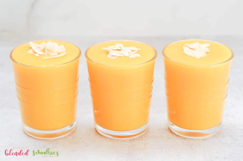 Three Smoothies Lined Up Next To Each Other - Mango Peach Smoothies With Coconut Flakes On Top In Clear Glasses