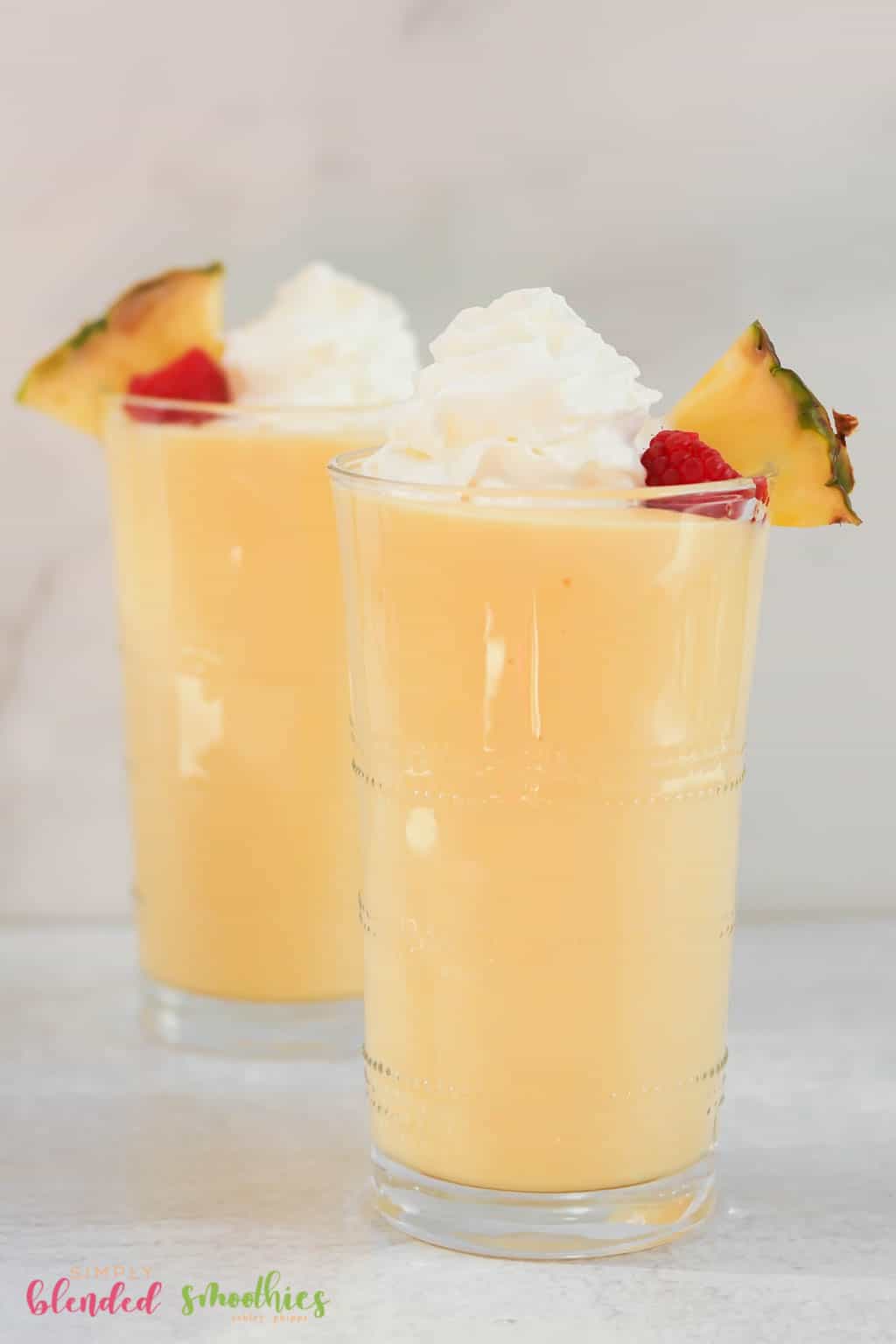 Vertical Photo Of Two Pineapple Peach Milkshakes Topped With A Pineapple Slice Raspberry And Whipped Cream